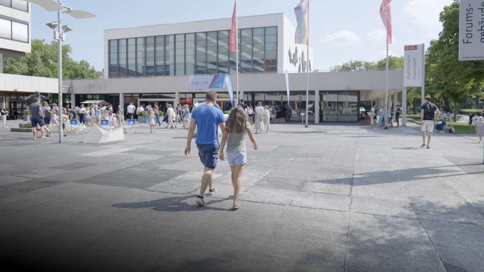 Picture of the forecourt of the AudiMax of the TU Braunscweig with play button to the video Checker Tobi live in the Audimax